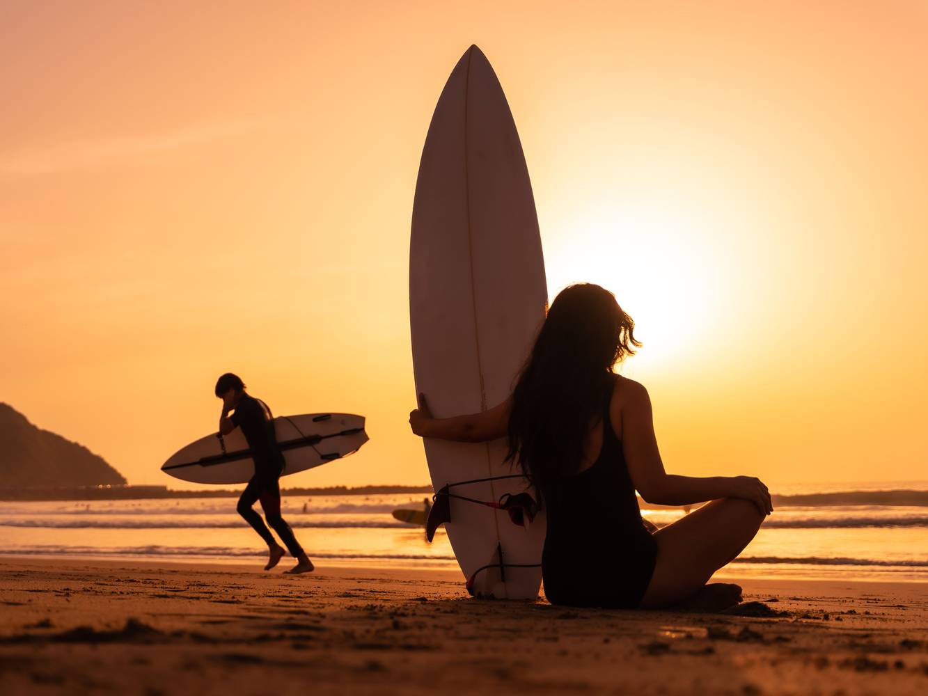 A Silhouette of a female from behind holding the surfboard with another surfer and a sunset view in the background